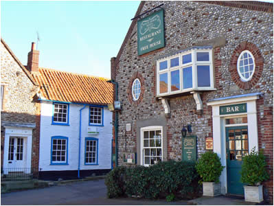 Stone facade of a quaint pub with a green sign, next to a white house with blue trim. Sunny day creates a welcoming, traditional village feel.