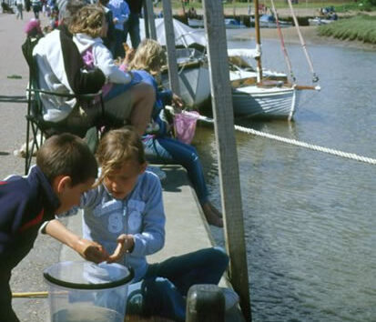 Children sitting on a riverside promenade, examining a net and bucket, with boats moored in the background. A relaxed, curious atmosphere.