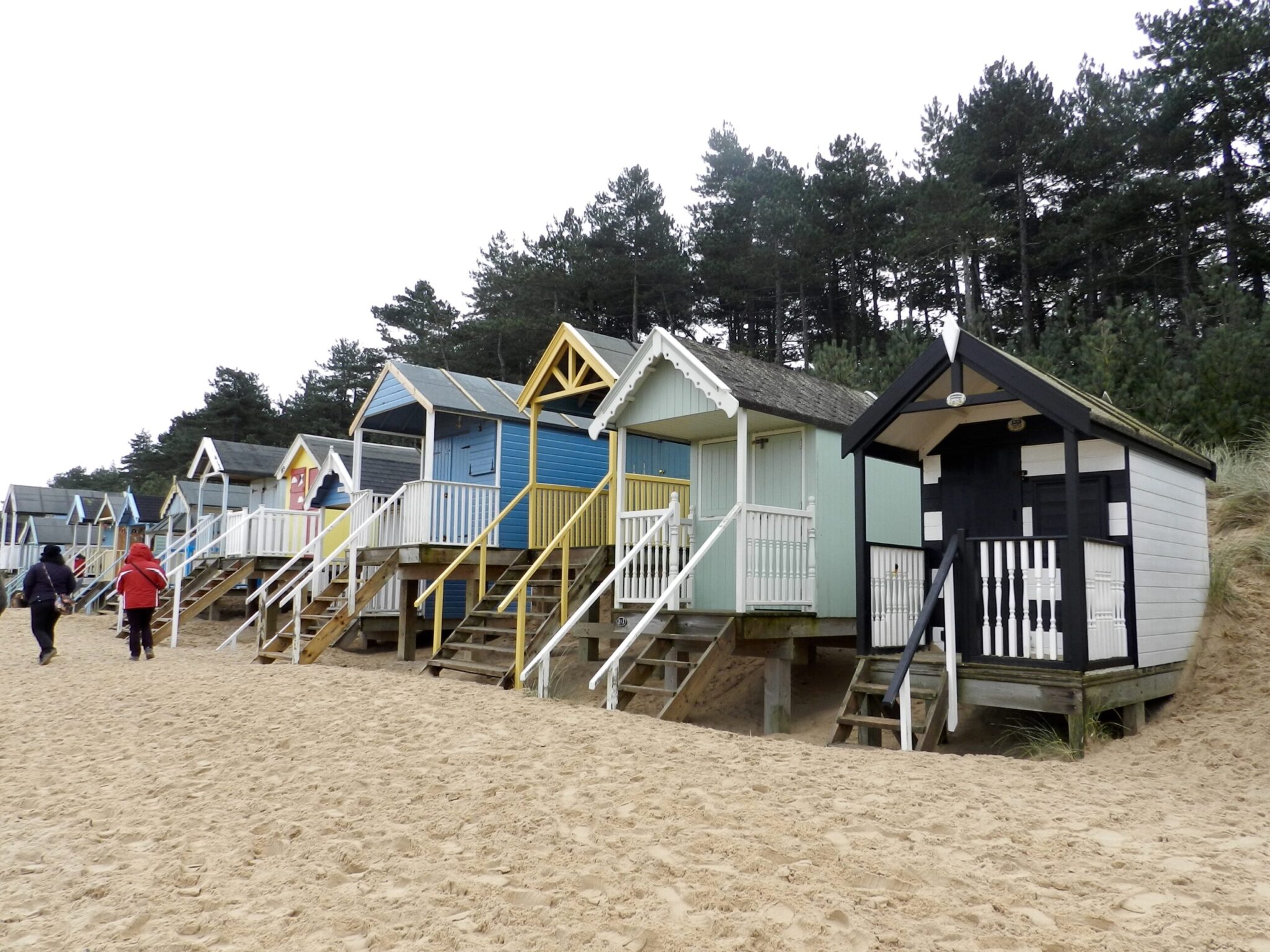 Colorful beach huts on sandy shores, surrounded by pine trees. People walk along the beach under overcast skies, creating a cozy, relaxed atmosphere.
