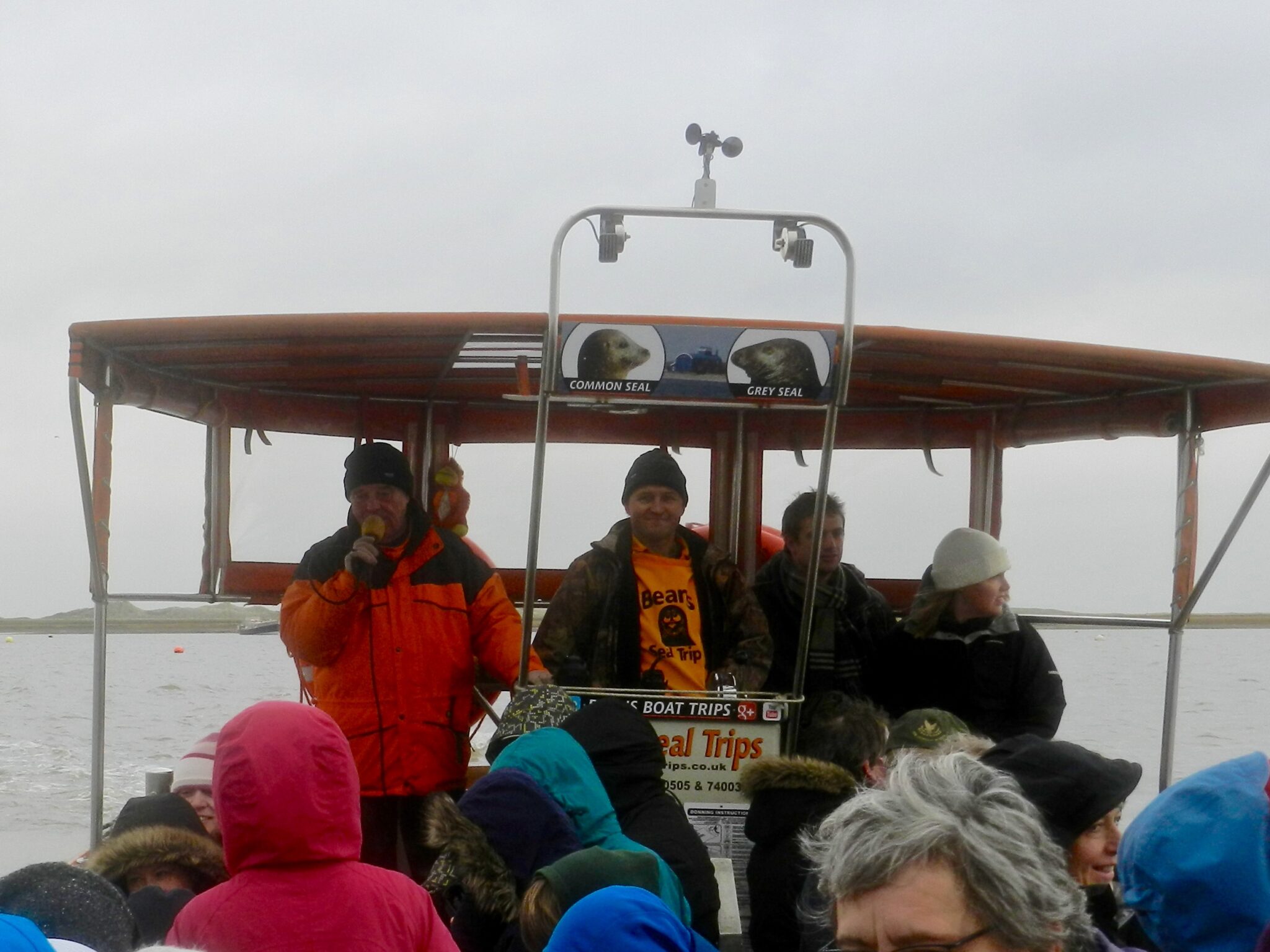 A group of people on a small boat with an orange canopy and signs for seal tours. Guides in bright jackets speak to bundled passengers. Overcast sky.