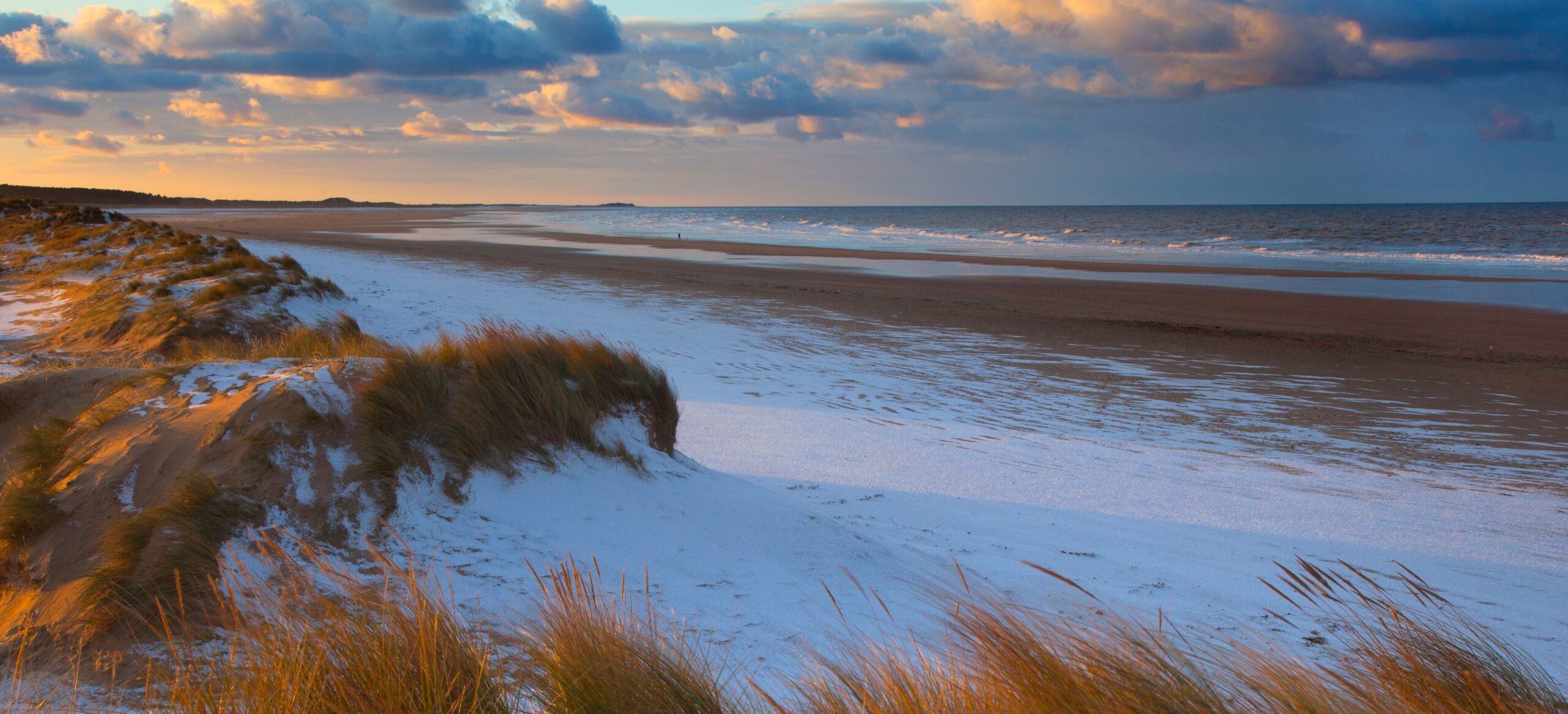 Coastal landscape at sunrise with grassy sand dunes in the foreground. A vast beach stretches to a calm ocean under a vibrant sky with scattered clouds.