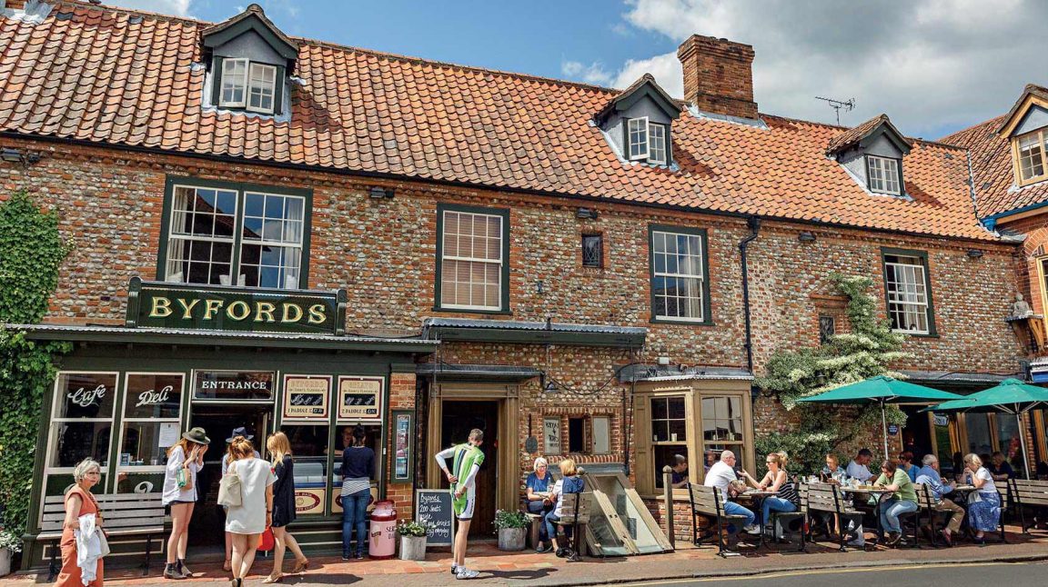 Brick cafe with a terracotta roof on a sunny day; people socialize outside under green umbrellas, creating a lively, relaxed atmosphere.