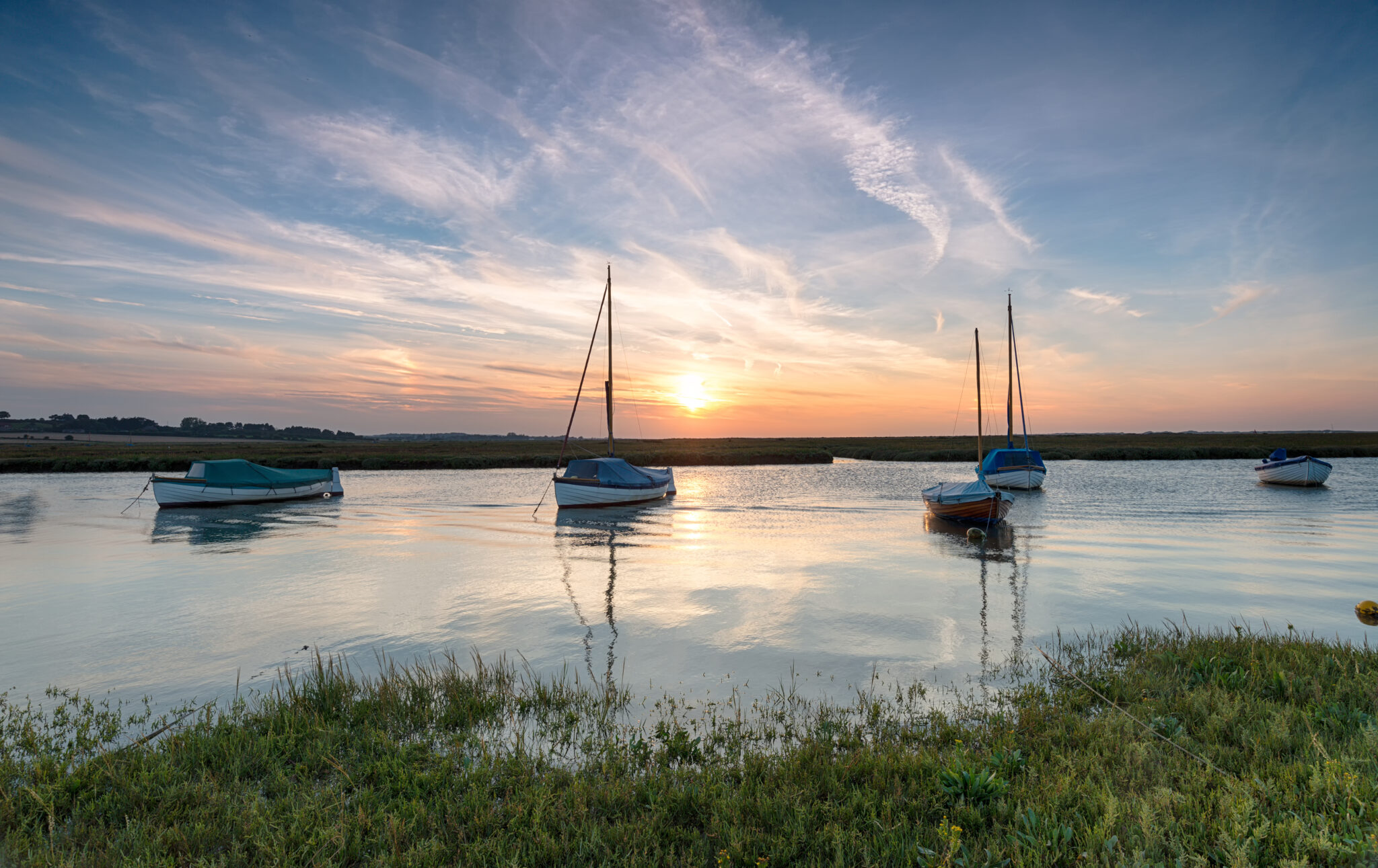 Boats peacefully anchored on calm water at sunset, with vibrant orange and blue skies reflecting on the water. Grassy shoreline in the foreground.
