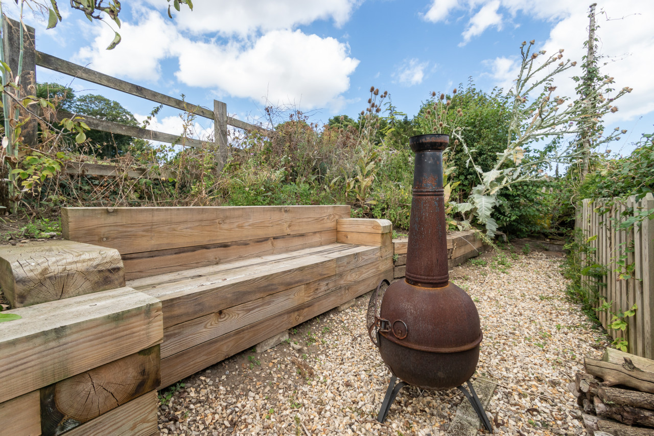 Wooden bench and rusted chiminea on a gravel path, surrounded by overgrown plants and a wooden fence, under a cloudy sky