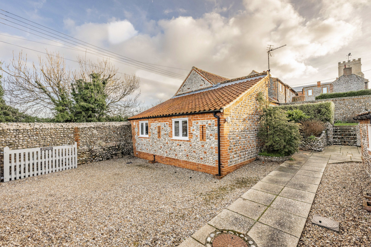 quaint cottage with a red-tiled roof and flint stone walls stands in a gravel courtyard. A white gate and stone path lead to steps, under a cloudy sky