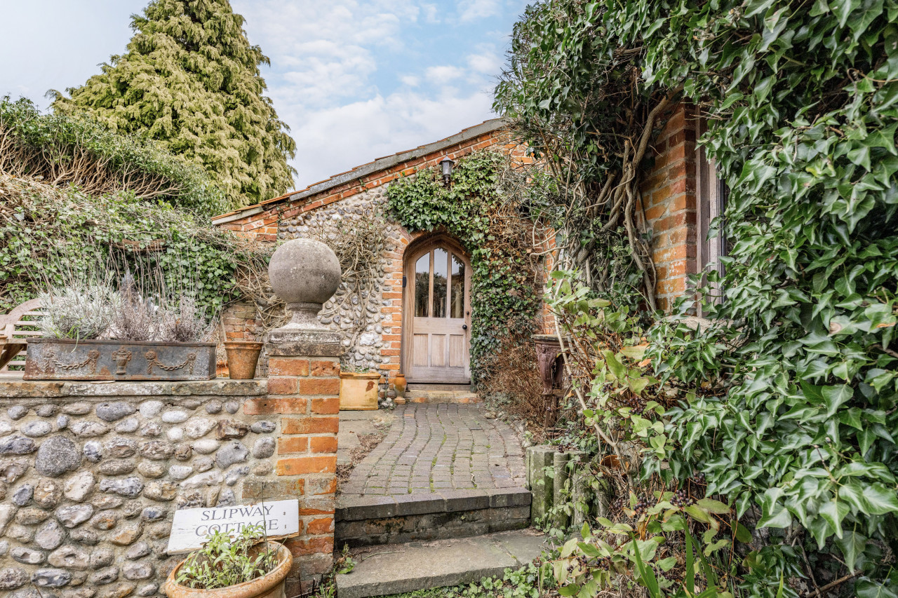 Charming brick and stone cottage entrance, framed by lush ivy and plants. A cobblestone path leads to an arched wooden door