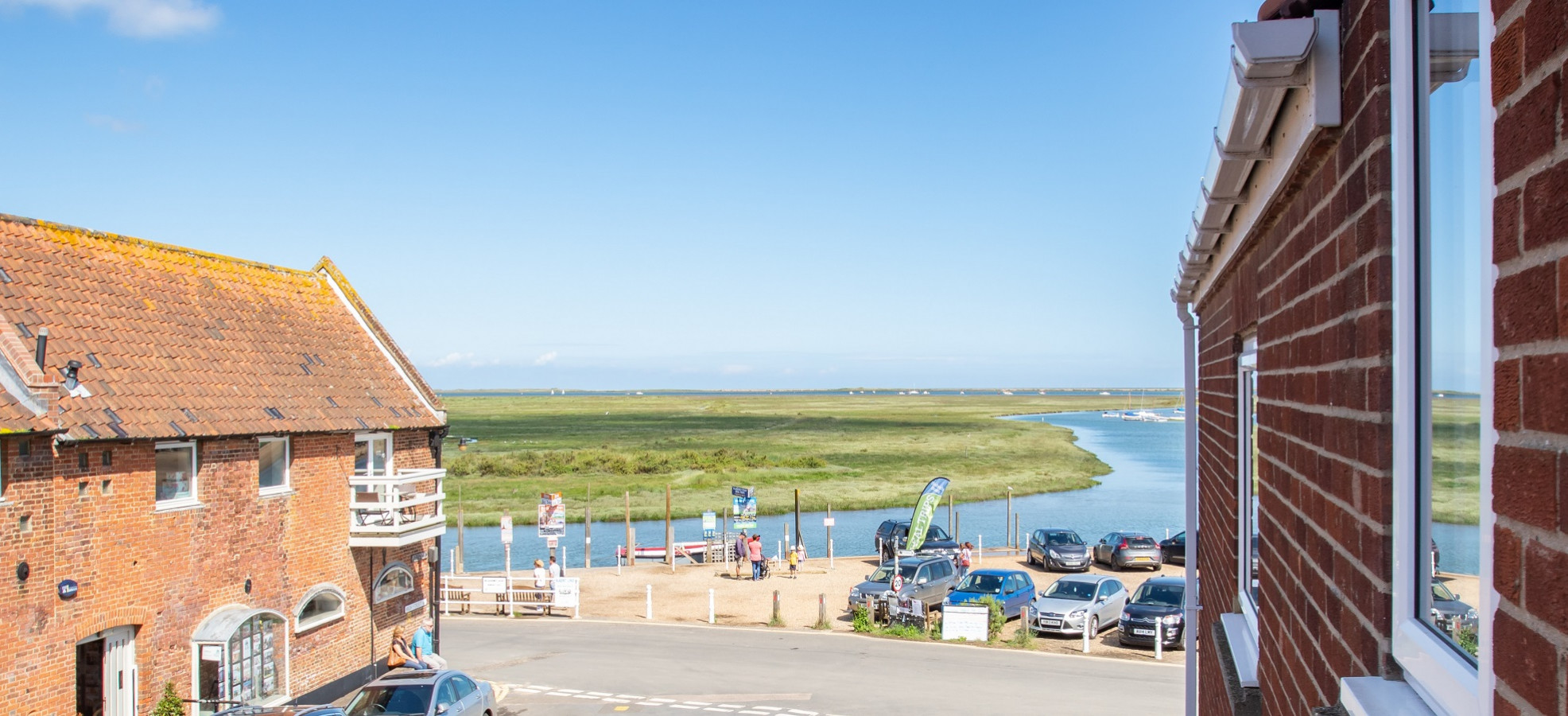 Coastal scene with a red brick building, parked cars, and people near a river under a clear blue sky. The river curves through green marshland, creating a peaceful atmosphere