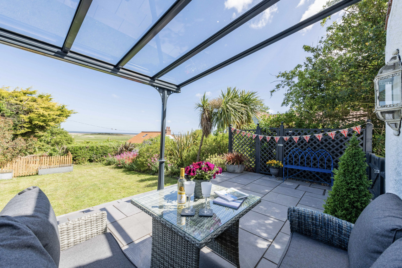 Covered patio with wicker seating and glass table, overlooking a lush garden with a distant sea view. Festive bunting and potted plants add a cheerful vibe