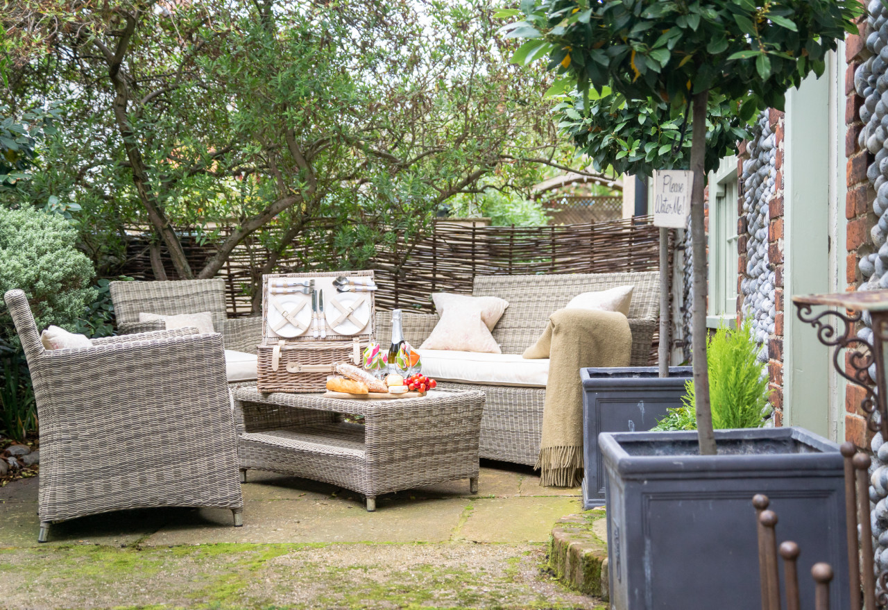 Outdoor patio with wicker furniture set amid lush greenery. A picnic basket, bread, and wine on the table create a cozy, inviting atmosphere