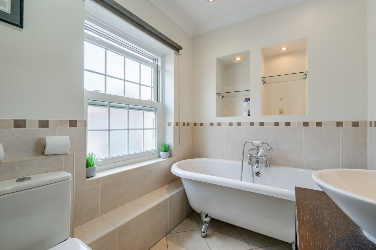 Bright bathroom with neutral tiles, featuring a clawfoot bathtub under a window with crossed panes. Shelves above hold toiletries; a serene, fresh vibe