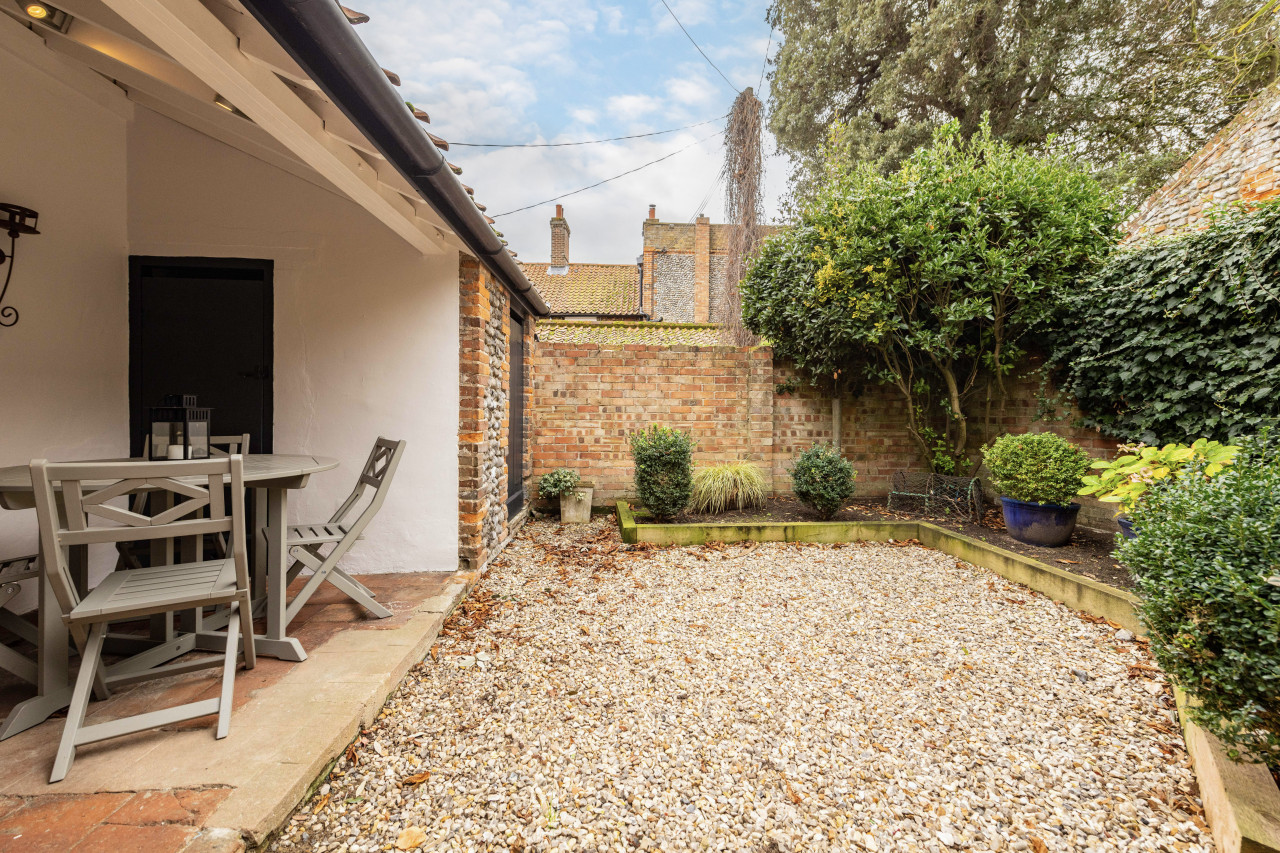 Cozy courtyard with gravel ground, bordered by brick walls and lush greenery. A patio set is to the left under a roofed section. Peaceful atmosphere