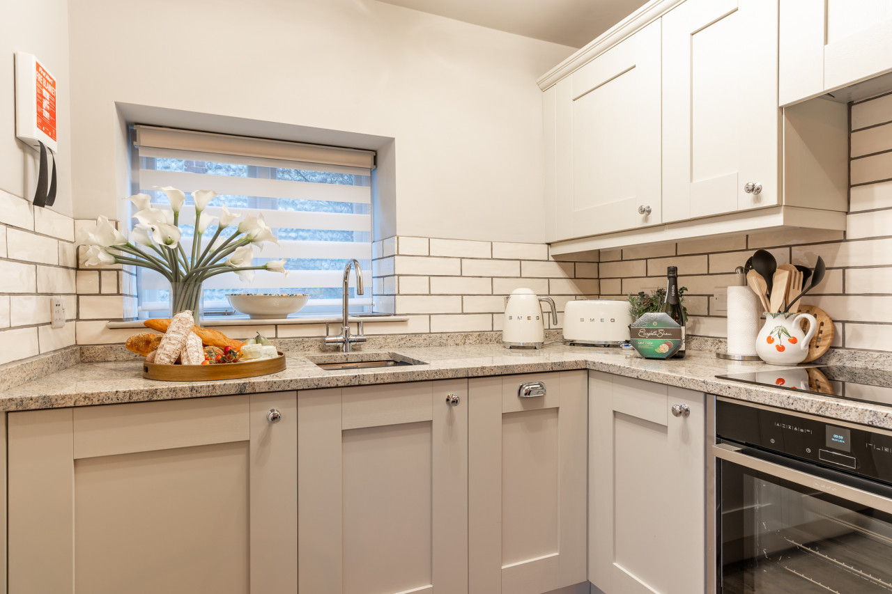 Cozy kitchen with light cabinets, granite countertops, and white brick backsplash. Features a Smeg toaster, kettle, fresh bread, flowers, and a wine bottle