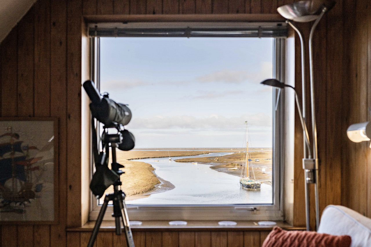 Cozy wooden room with a telescope aimed at a large window view of a serene river estuary. A sailboat is docked, evoking tranquility and exploration