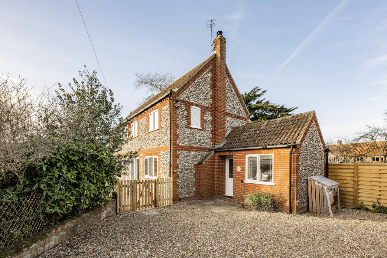 uaint stone and brick house with a pitched roof, surrounded by greenery. A gravel driveway leads to a wooden picket gate. The setting is peaceful