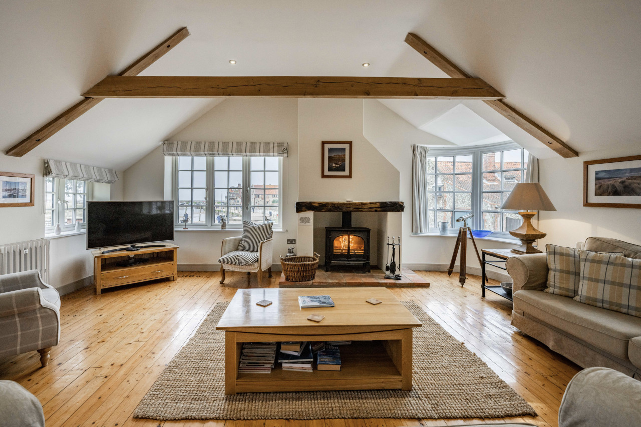 Cozy living room with wooden beams, a warm fireplace, and soft lighting. Includes comfy sofas, a coffee table on a rug, and a TV. Relaxing atmosphere
