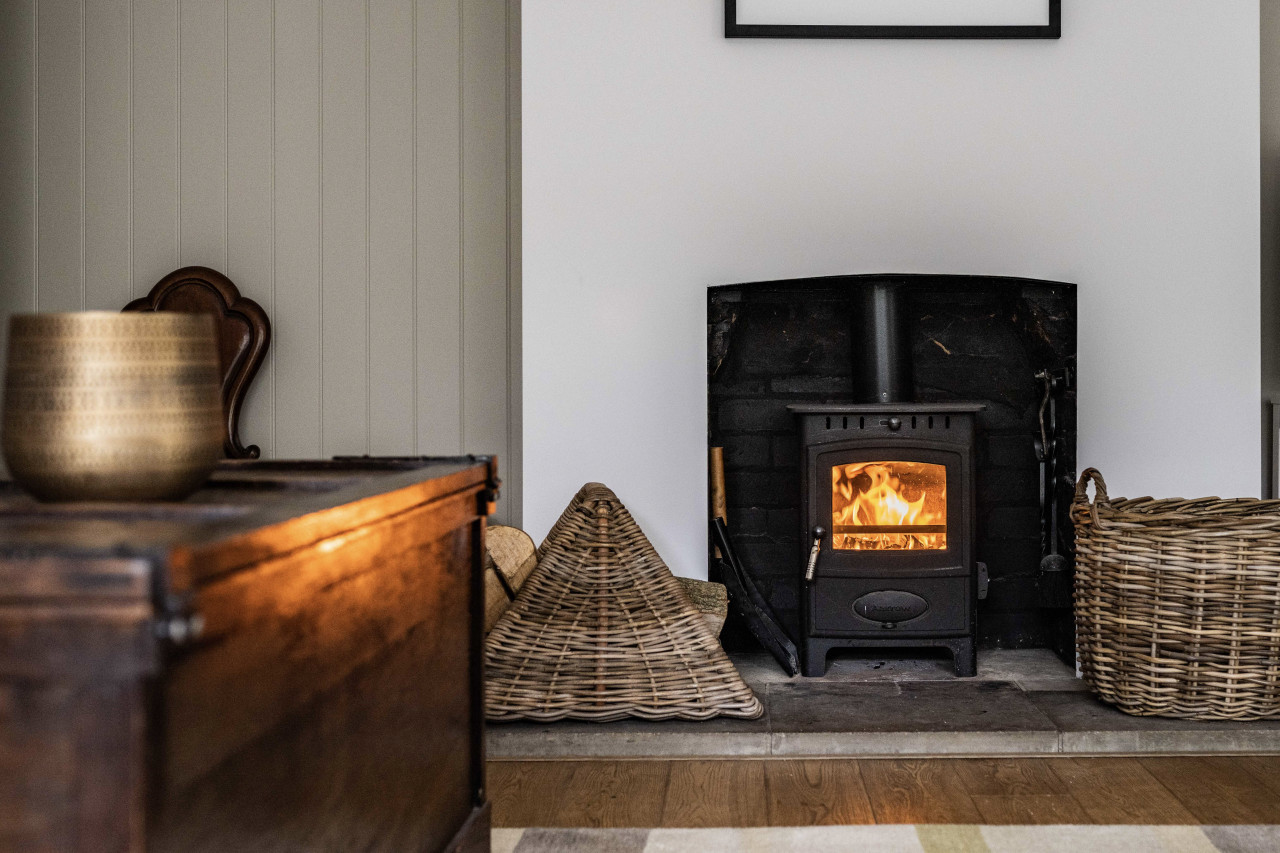 Cozy room with a warm, lit fireplace in a black stove, flanked by wicker baskets. A wooden chest and vase enhance the rustic ambiance.