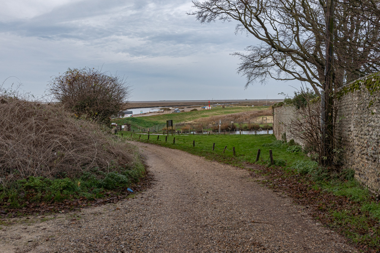 winding dirt path leads through a rural landscape with fields, trees, and a stone wall under a cloudy sky, conveying a calm, serene atmosphere