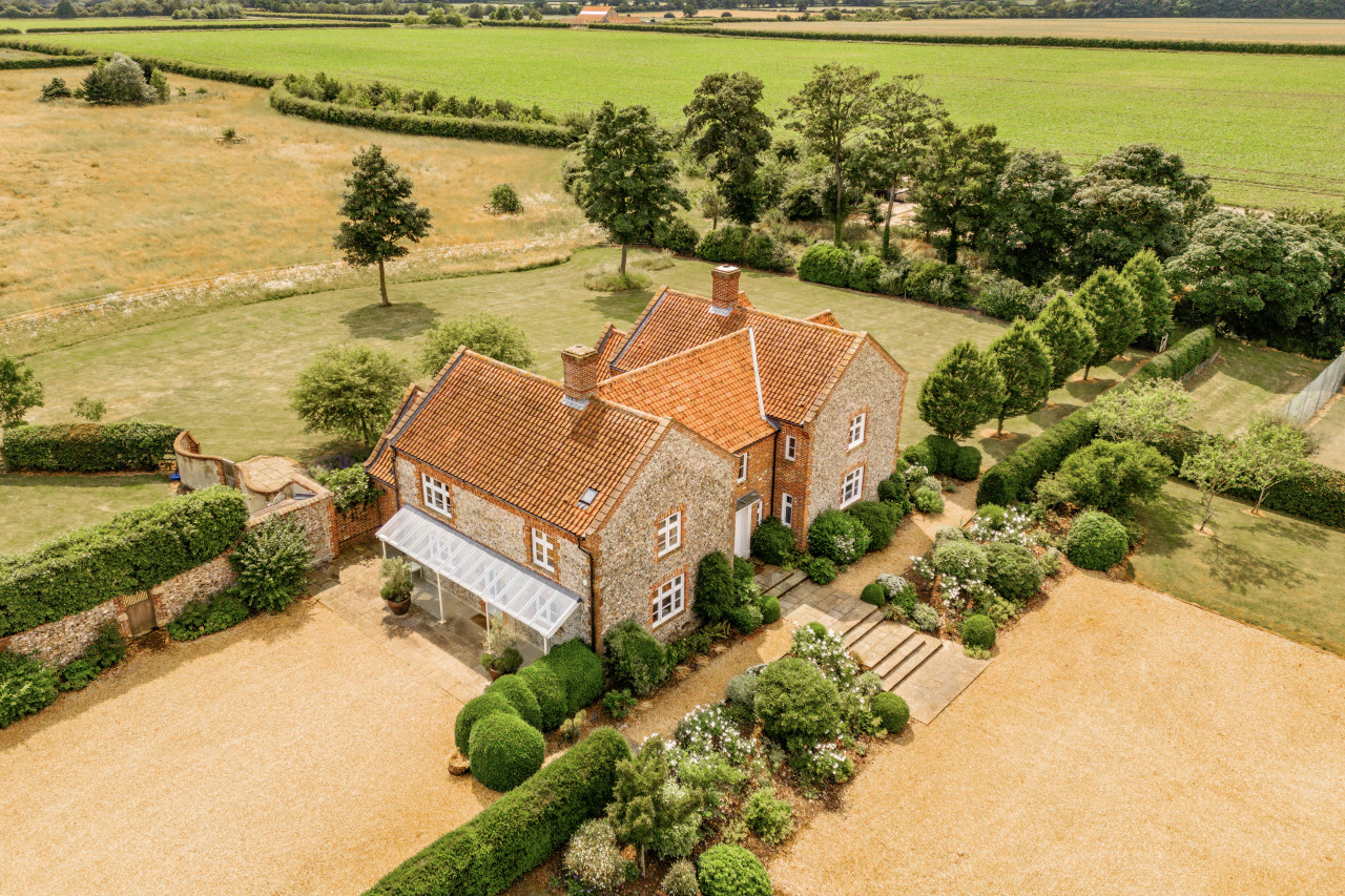 Aerial view of a charming countryside house with red tile roof, surrounded by lush greenery and gardens. Vast fields stretch into the distance, exuding tranquility