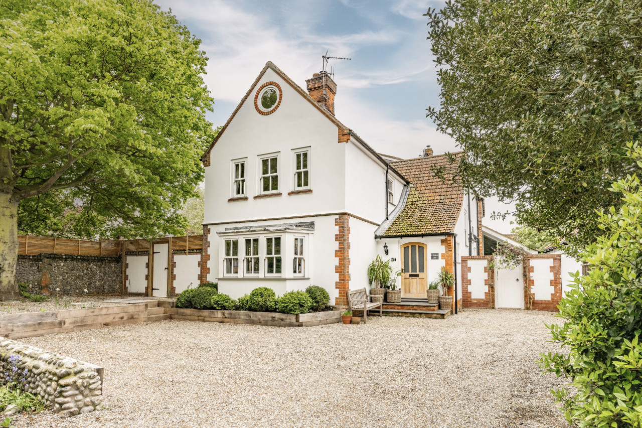 White cottage with red brick accents, surrounded by lush trees. Gravel driveway leads to wooden door, with potted plants and a bench. Tranquil ambiance