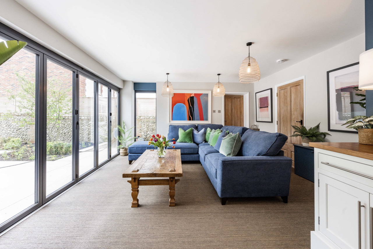 Modern living room with blue sectional sofa adorned with green pillows, wooden coffee table with tulips, and floor-to-ceiling windows on the left