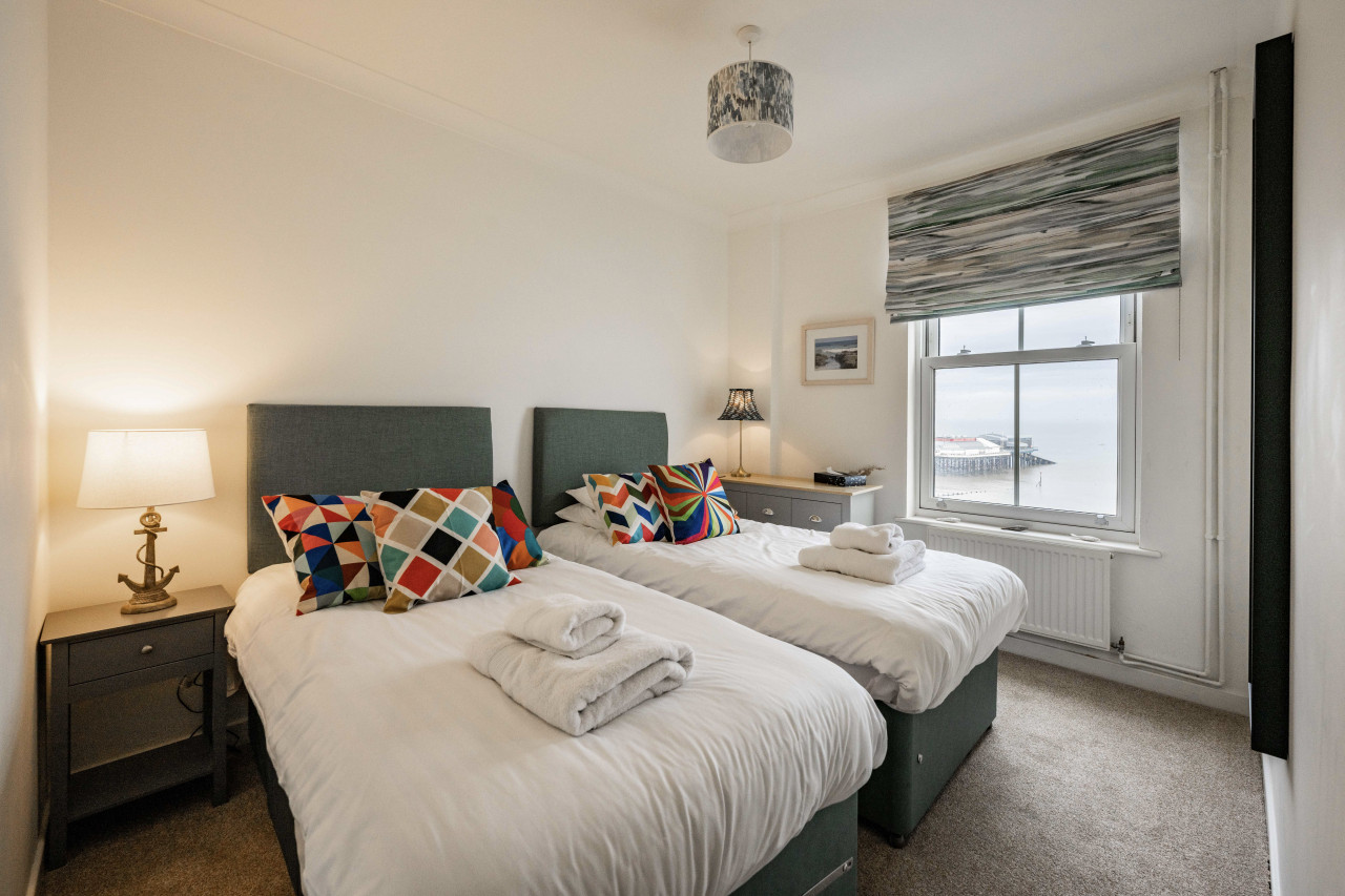 Two neatly made single beds with colorful geometric cushions in a cozy room. A window reveals a seaside view. Soft lighting gives a warm, inviting tone