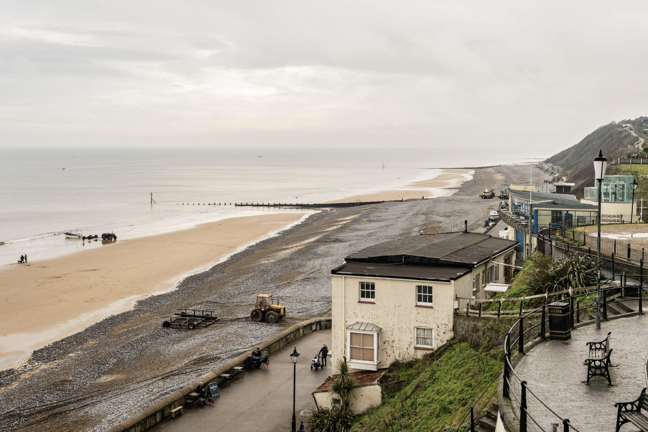 Coastal scene with overcast skies, sandy beach, and a pebble path. A lone tractor is parked near a weathered building, conveying a calm, serene mood