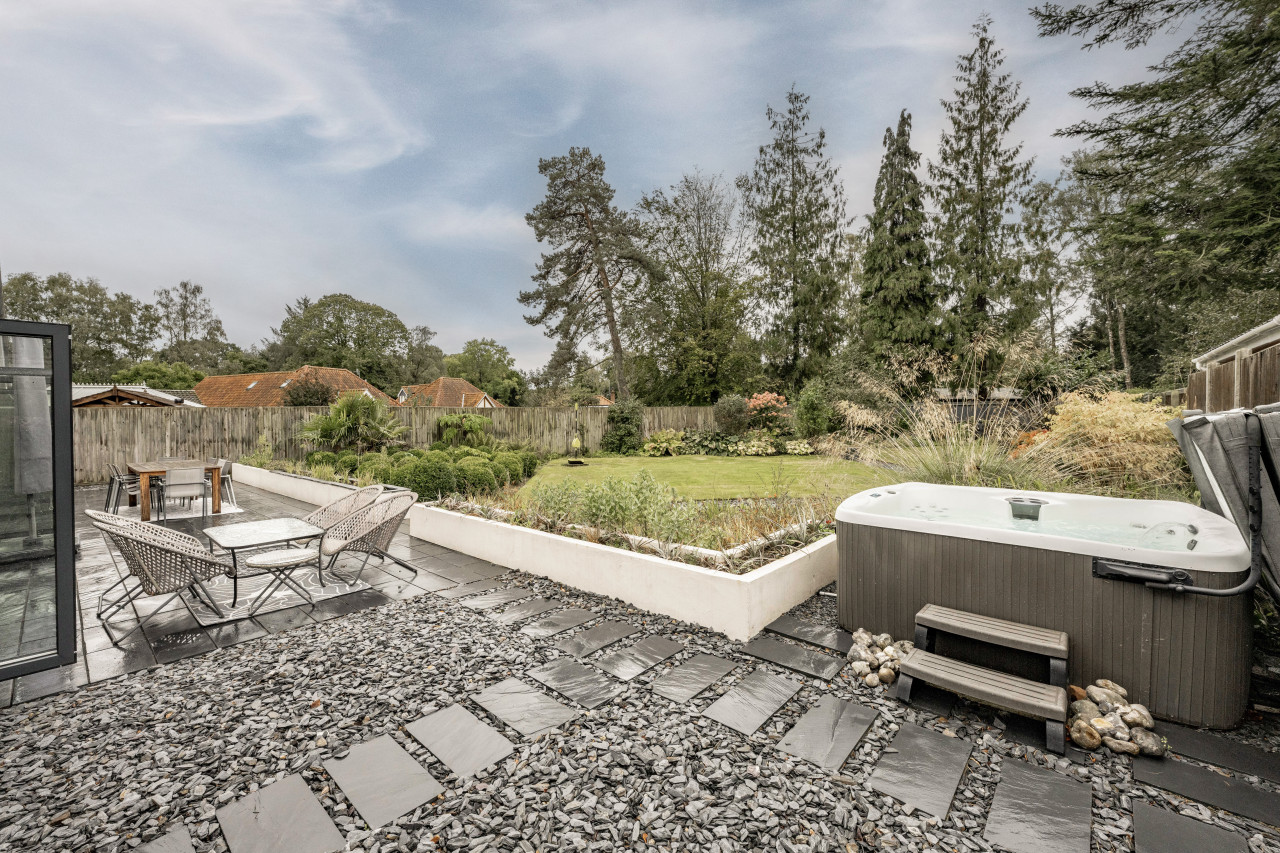 Spacious backyard with a hot tub on a stone patio, surrounded by lush greenery and trees. A dining set and a pathway enhance the tranquil outdoor setting