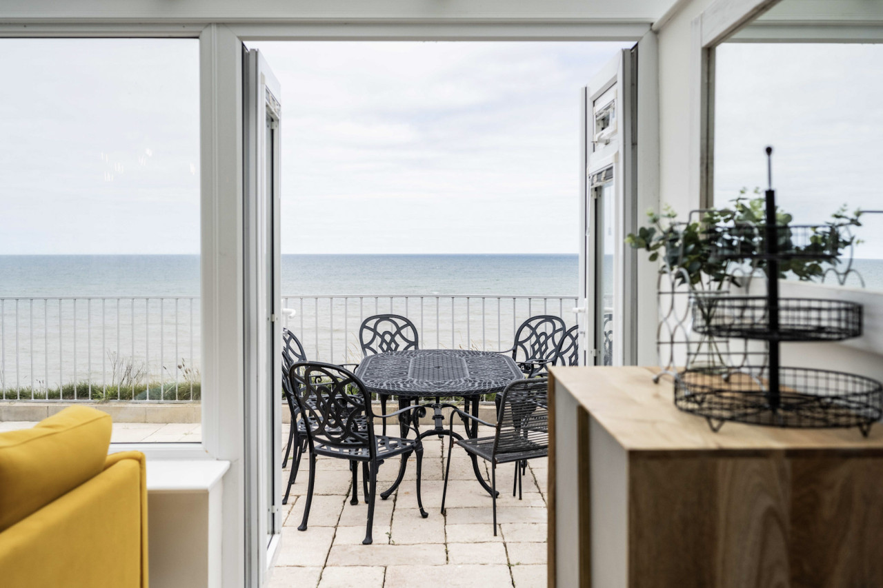 Bright coastal room with open doors leading to a patio; features elegant black wrought iron table and chairs, ocean view, and yellow sofa accent