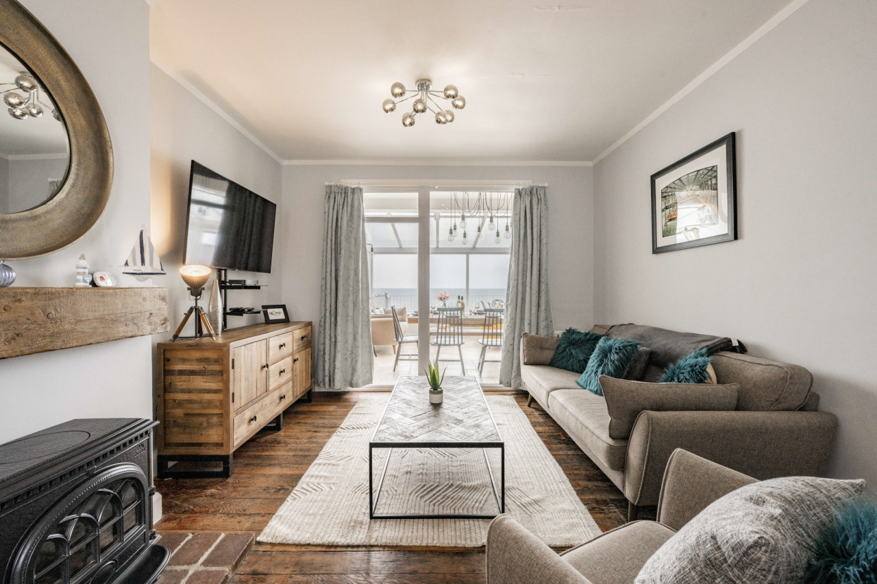 Modern living room with a cozy atmosphere, featuring a beige sofa with teal cushions, a large TV, stylish wooden furniture, and a sea view through glass doors.