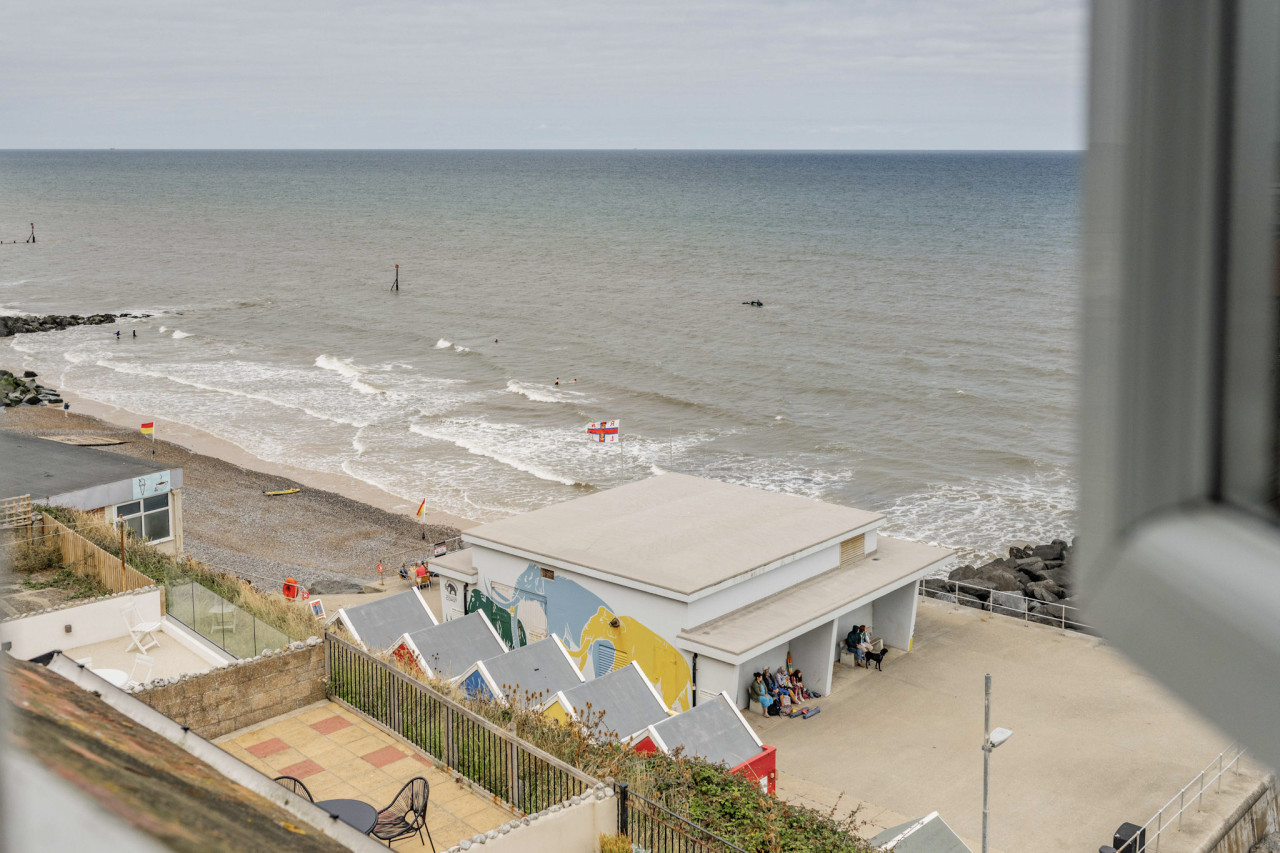 View from above of a sandy beach with gentle waves. A group of people relax near colorful beach huts. The scene is calm and overcast