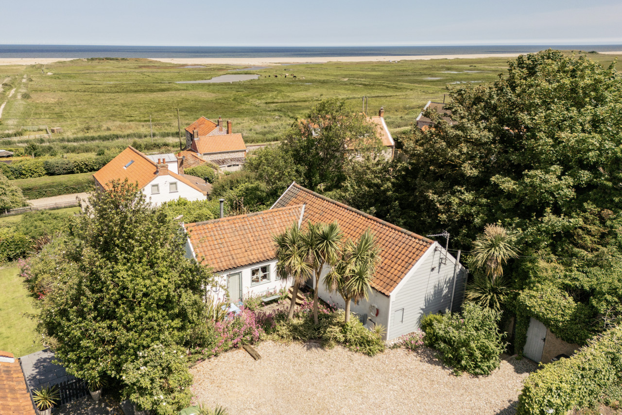 Aerial view of a quaint coastal cottage with a red tile roof, surrounded by lush greenery and palm trees. The sea and grassy dunes appear in the distance