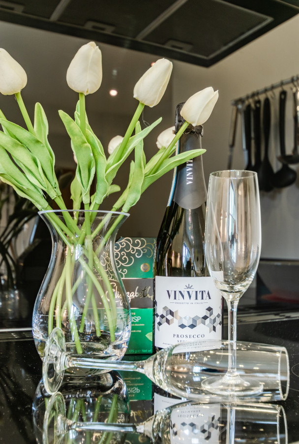 A vase of white tulips beside a bottle of prosecco and two champagne flutes on a glossy countertop, conveying a sense of elegance and celebration.