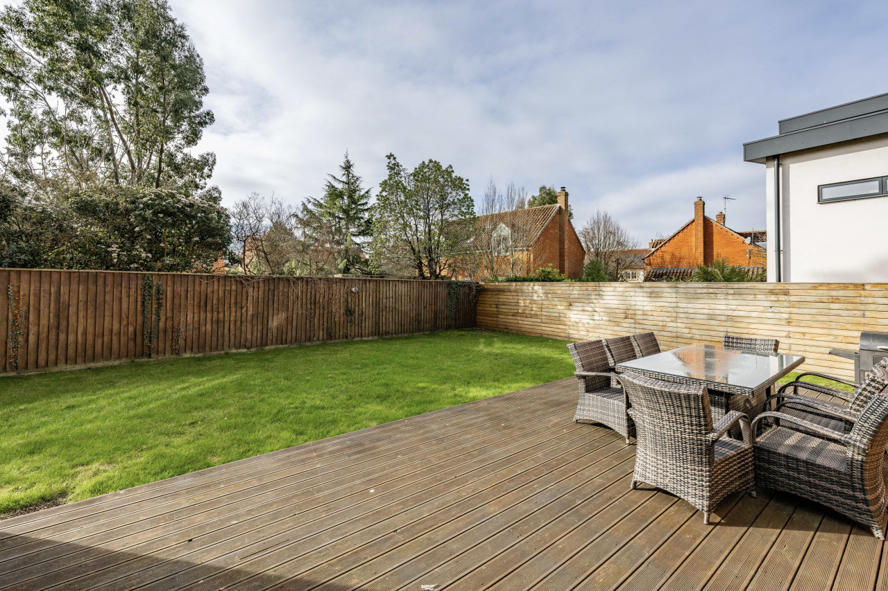 Spacious backyard with wooden deck, wicker dining set, and lush green lawn. High wooden fence and red brick houses under a partly cloudy sky.
