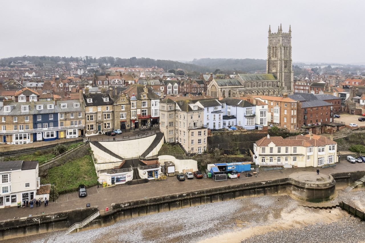 Aerial view of a coastal town with colorful historic buildings along the shore, a prominent stone church tower in the background, and a sandy beach.