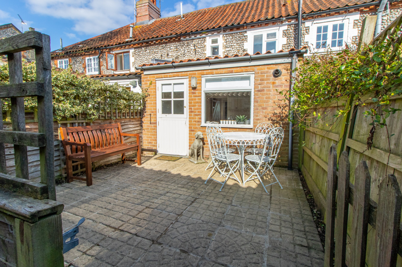 Charming backyard patio with brick walls and red tiled roof. Features a wooden bench, metal table with chairs, dog statue, and leafy fence. Sunny and inviting.