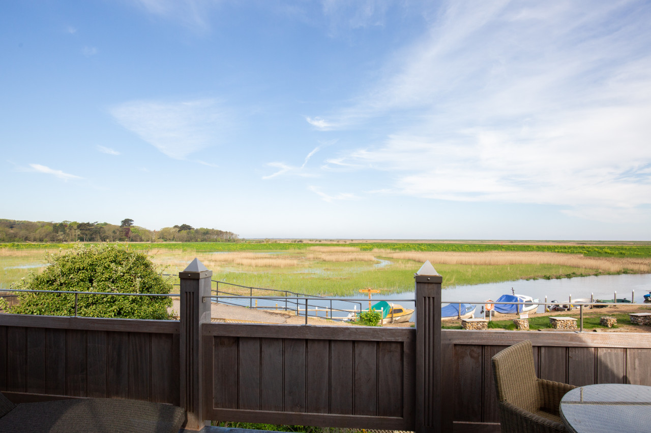Scenic view of a marshland under a clear blue sky, seen from behind a wooden fence. A winding river and small boats add serenity to the scene.