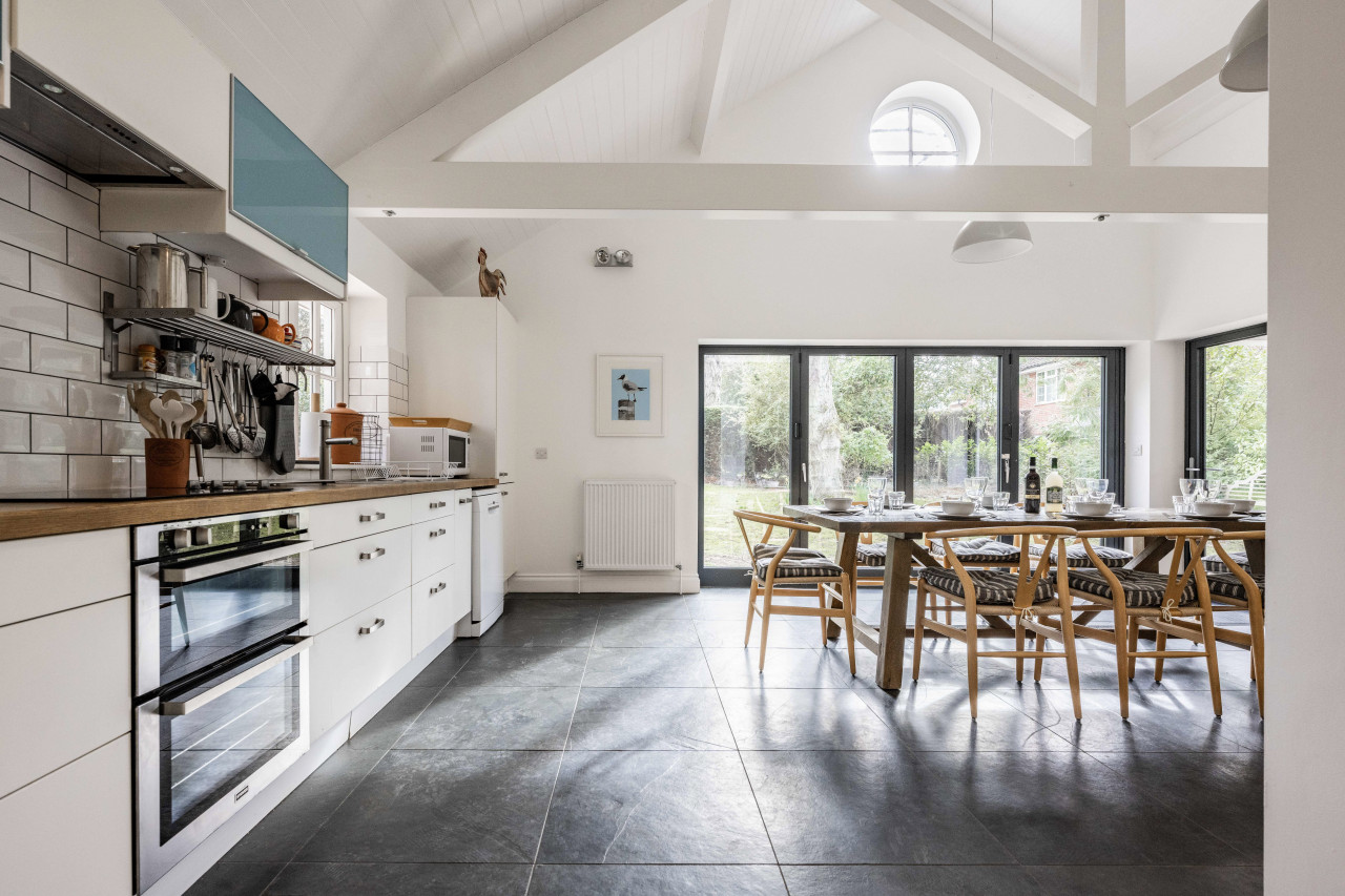 Spacious kitchen with white cabinets, wooden countertops, and a dining area. Sunlit through large windows, creating an airy, inviting atmosphere.