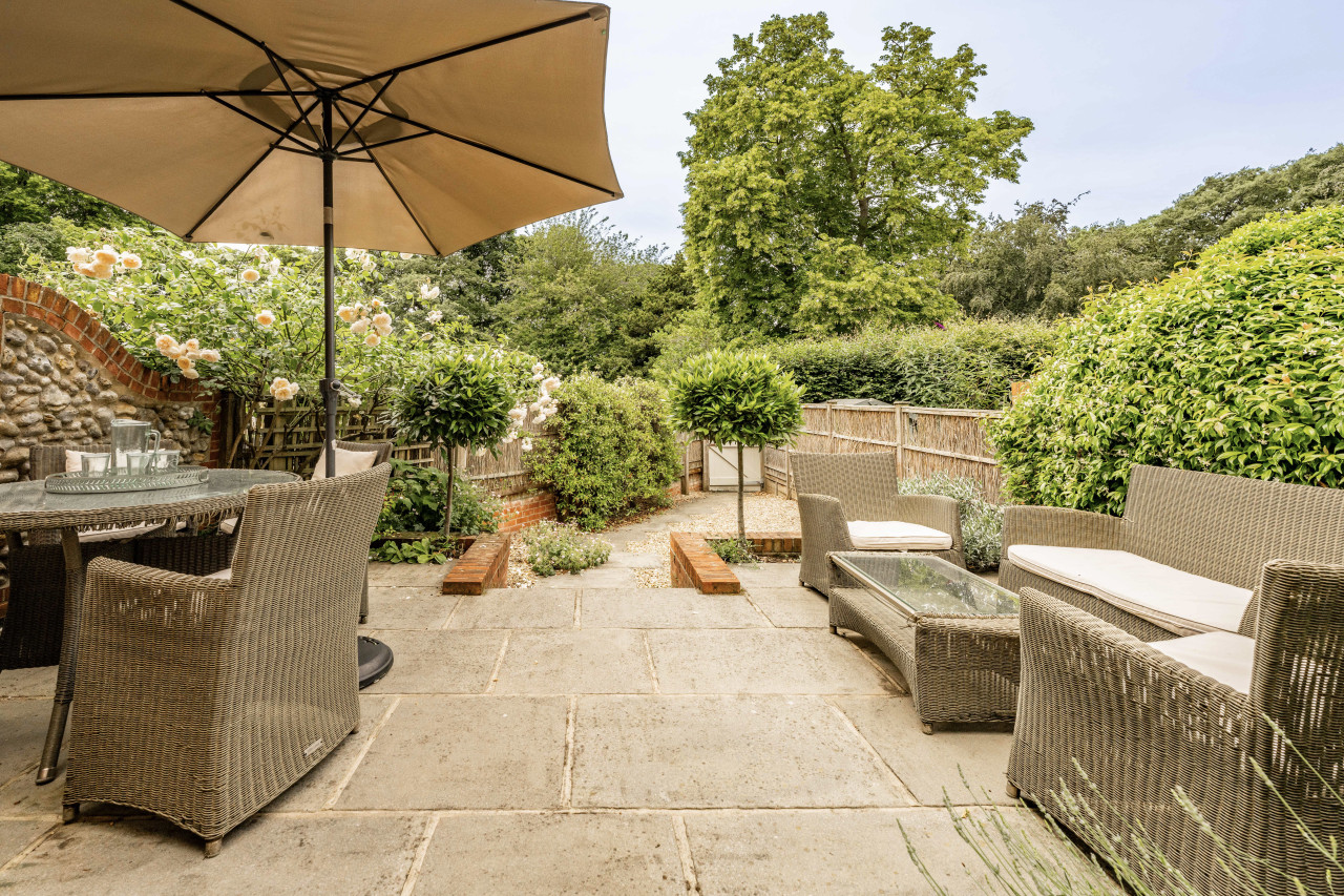 Outdoor patio with wicker furniture, glass table, and large umbrella. Surrounded by blooming flowers, lush greenery, and a wooden fence. Peaceful and inviting.