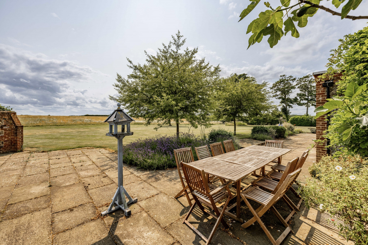 A tranquil garden patio features a long wooden table with chairs, surrounded by greenery. A birdhouse stands nearby under a partly cloudy sky.