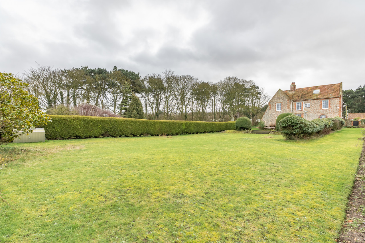 Large green lawn with a traditional brick house on the right, surrounded by hedges and trees under a cloudy sky, conveying a serene rural atmosphere.