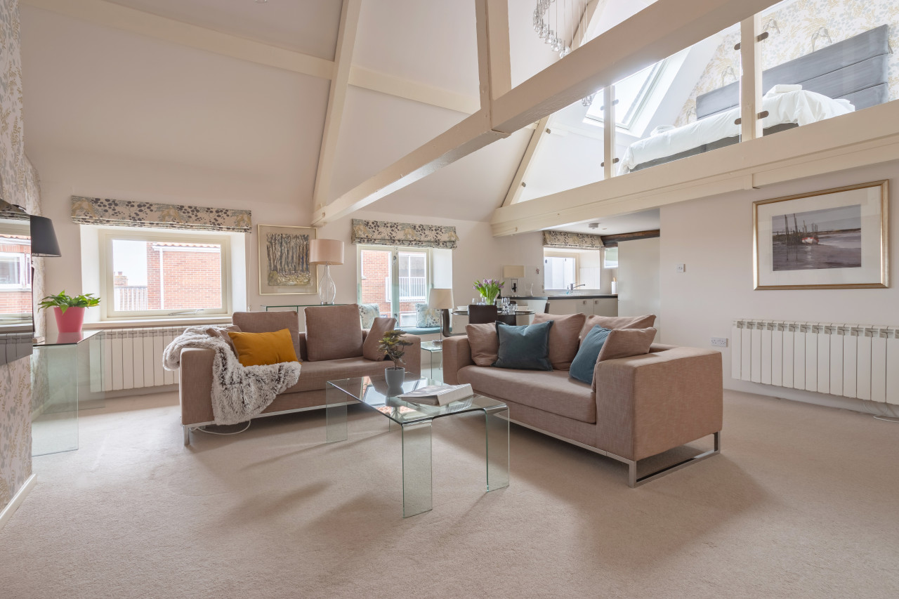 Spacious loft living room with beige sofas adorned with colorful cushions, glass coffee tables, exposed beams, soft lighting, and large skylight.