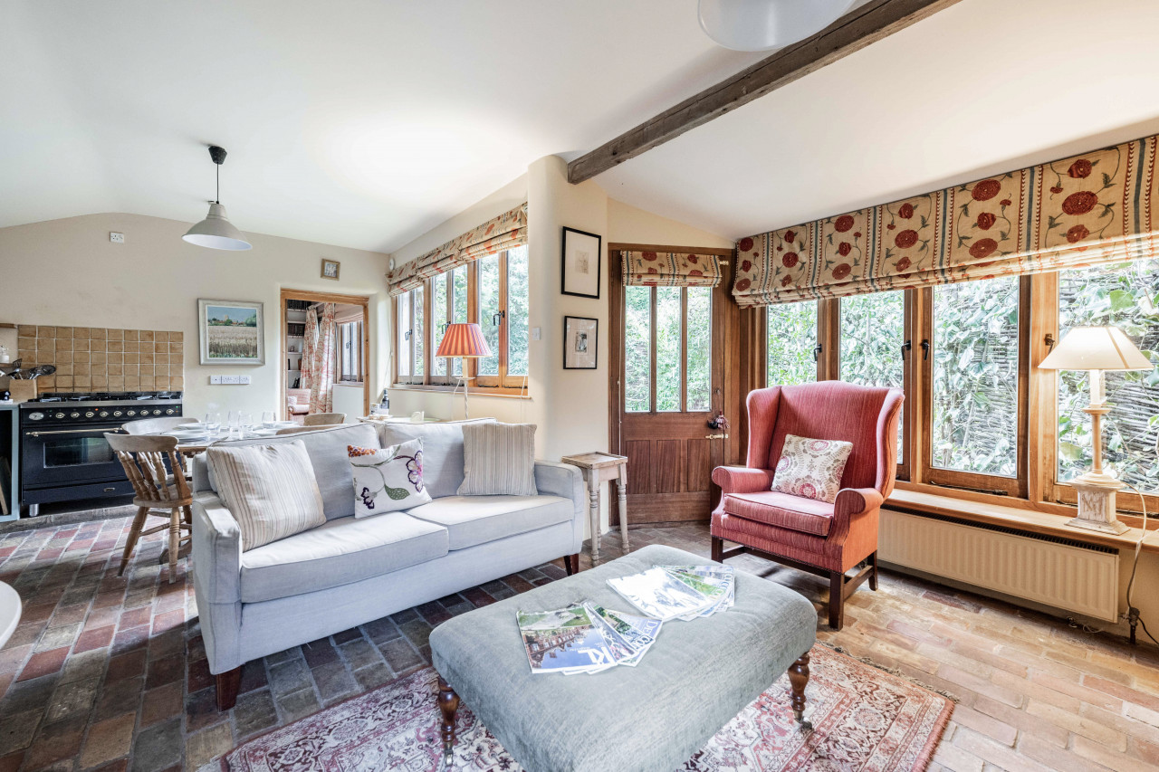 Cozy living room with brick flooring, featuring a light gray sofa, red armchair, and patterned curtains. Sunlit windows and warm, inviting decor.