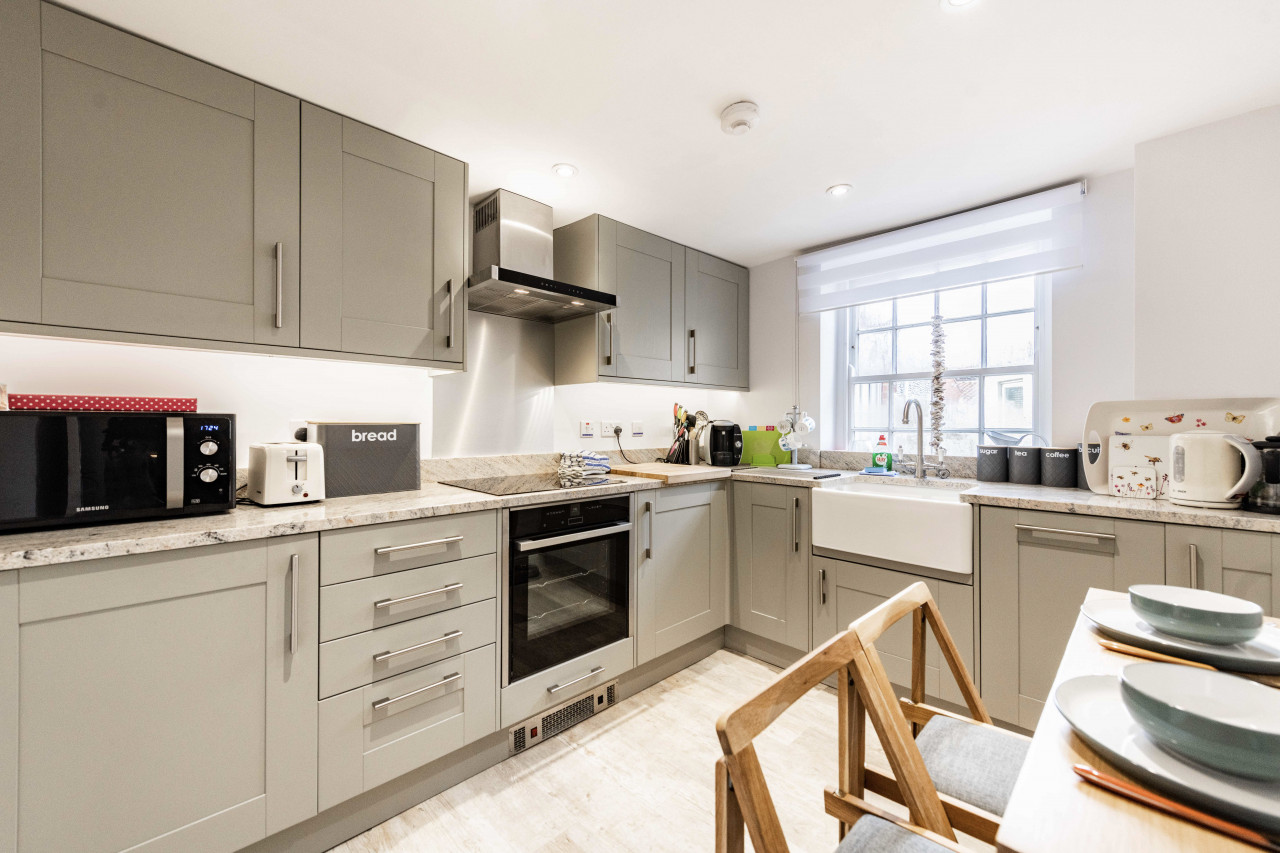 Modern kitchen with grey cabinets, a marble countertop, and stainless steel appliances including an oven and microwave. Wooden chairs and white crockery add a cozy touch.