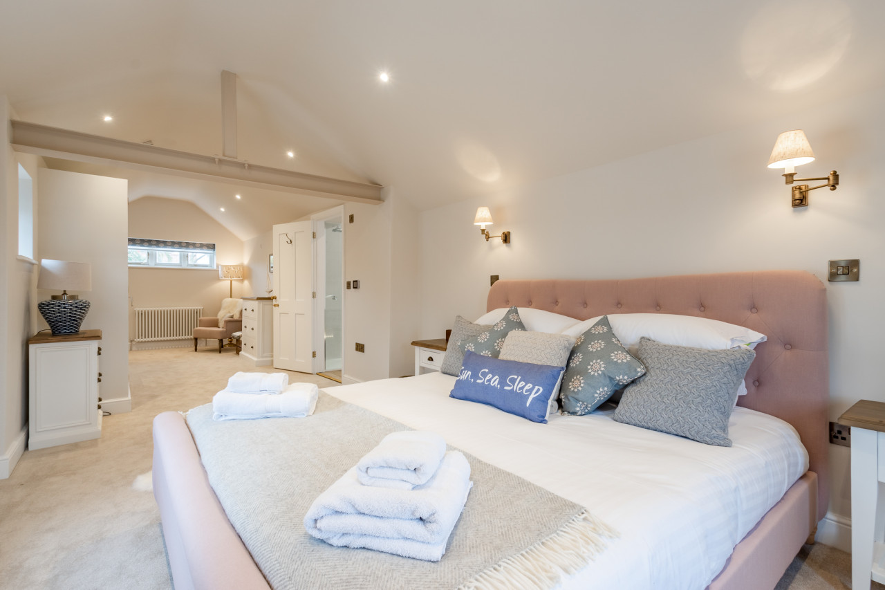 A cozy, elegant bedroom features a plush pink bed with a "Sun, Sea, Sleep" pillow. Soft lighting, neutral tones, and fluffy towels create a serene, welcoming ambiance.