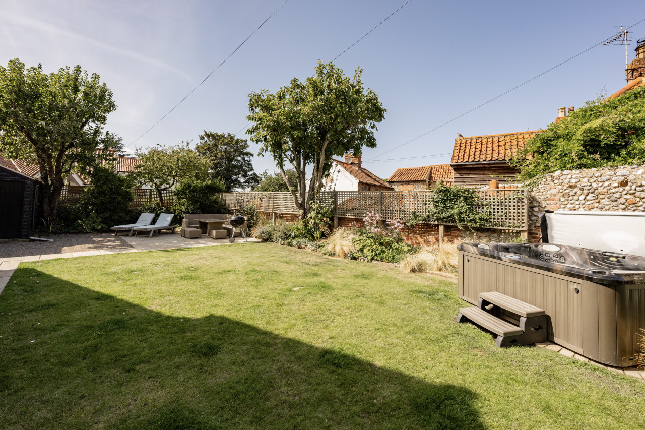 Sunny backyard with green lawn, hot tub, and patio furniture. Surrounded by trees and a stone wall with lattice, it feels peaceful and inviting.